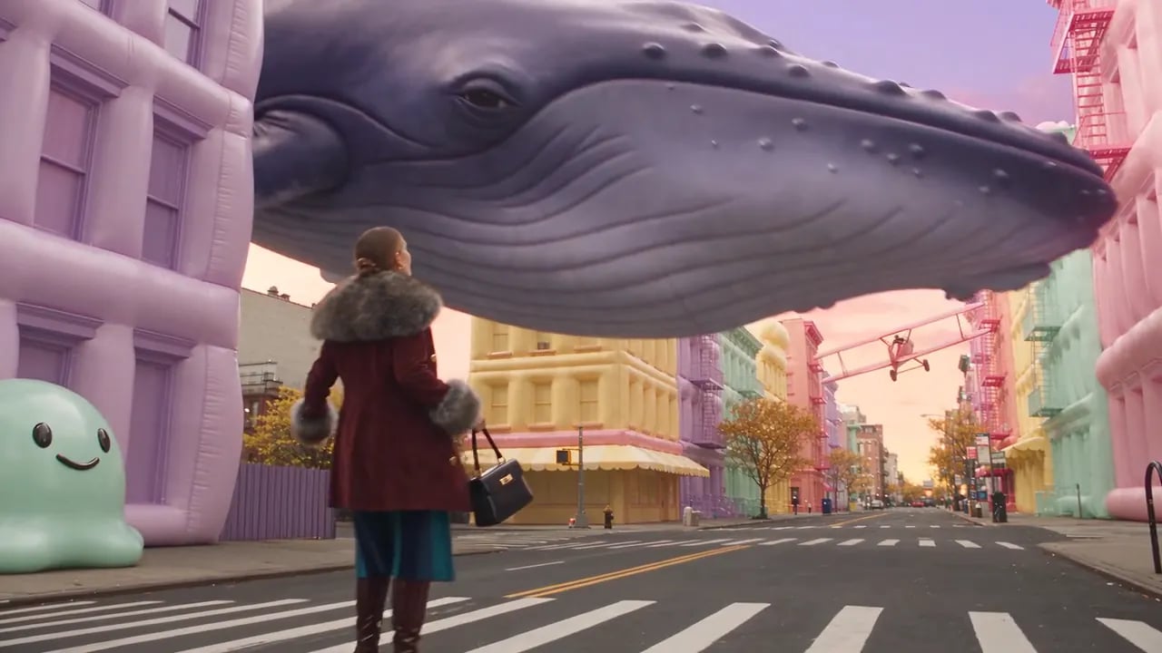 A woman in a fur-trimmed coat walks down a New York street that has turned into a pastel city with inflatable taxis and a floating whale.