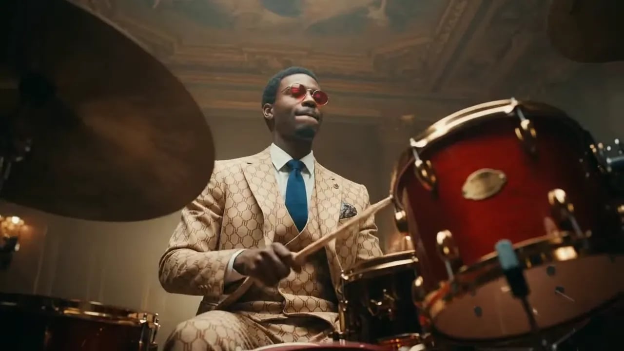 A drummer in a beige suit plays a red drum kit before the camera whip-pans to saxophonists and a ballet dancer under purple lights.