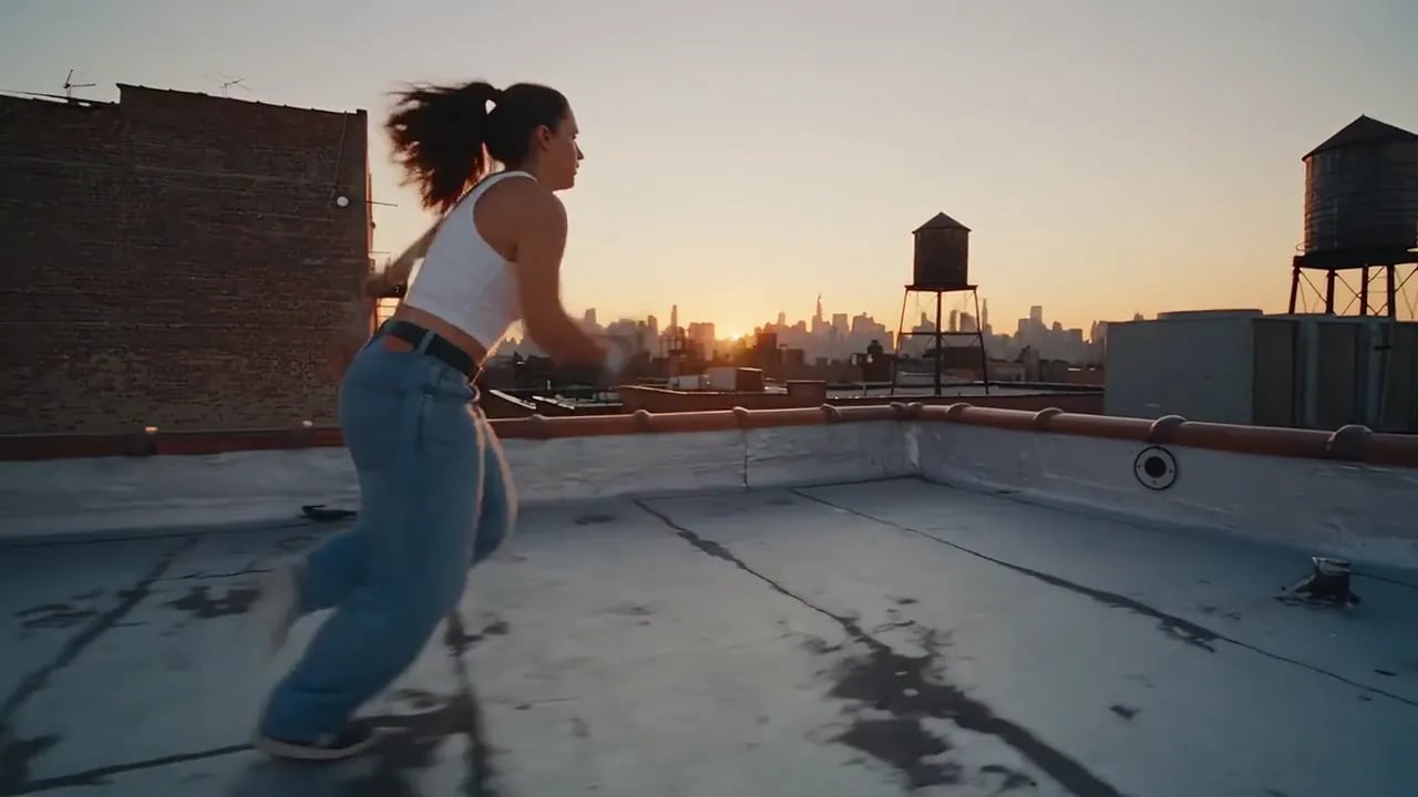A young woman in a white top and jeans runs and leaps across city rooftops at sunset.