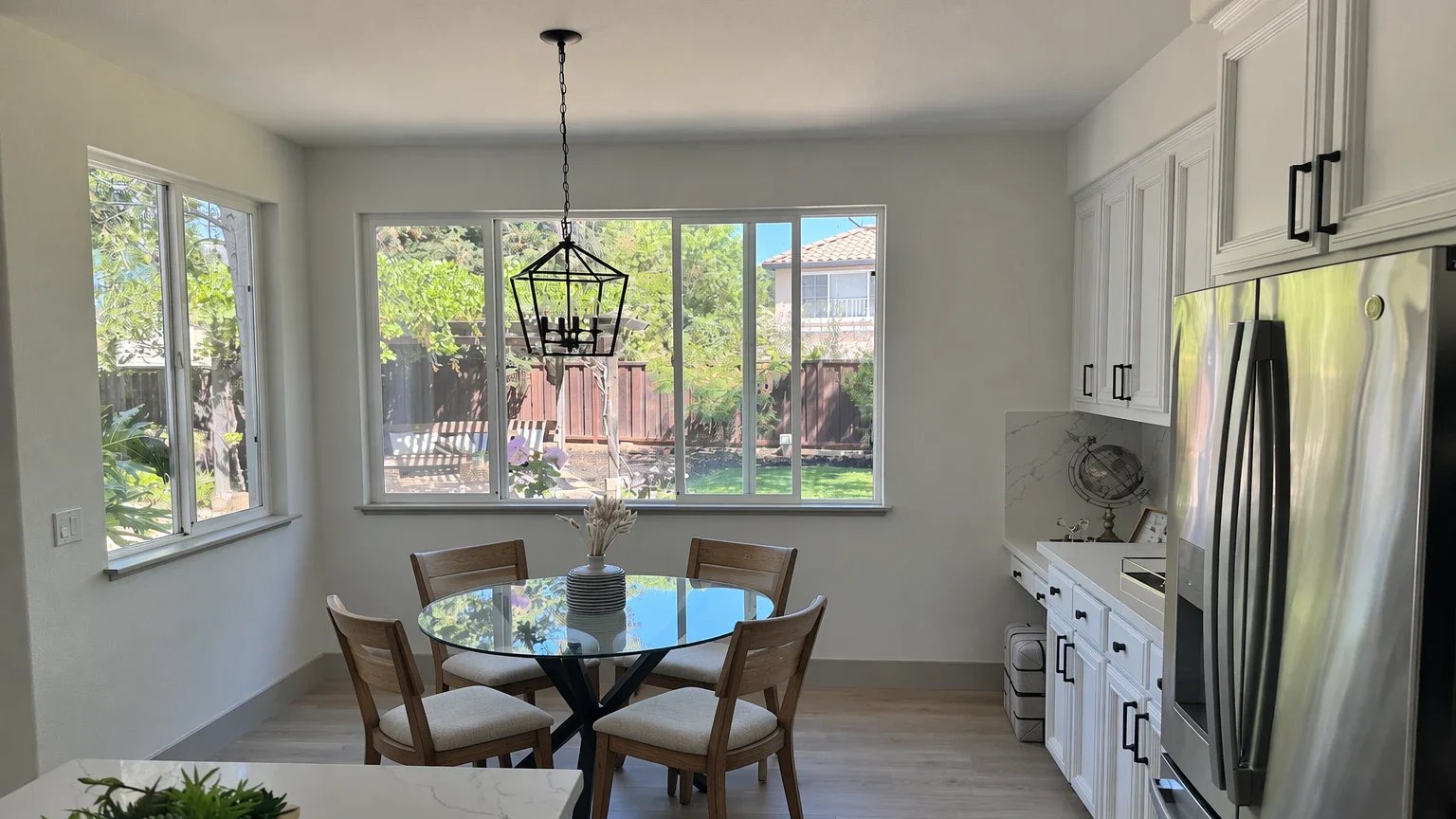 GPT Image 2.5 Flare edit of a bright dining nook: the white chairs are replaced with slatted wooden chairs with cream seats, and the glass table, pendant lamp and windows stay as they were.