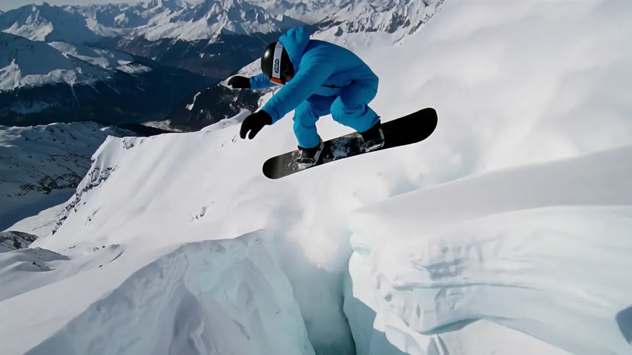 A snowboarder in a blue suit jumps a glacier crevasse on a steep slope.