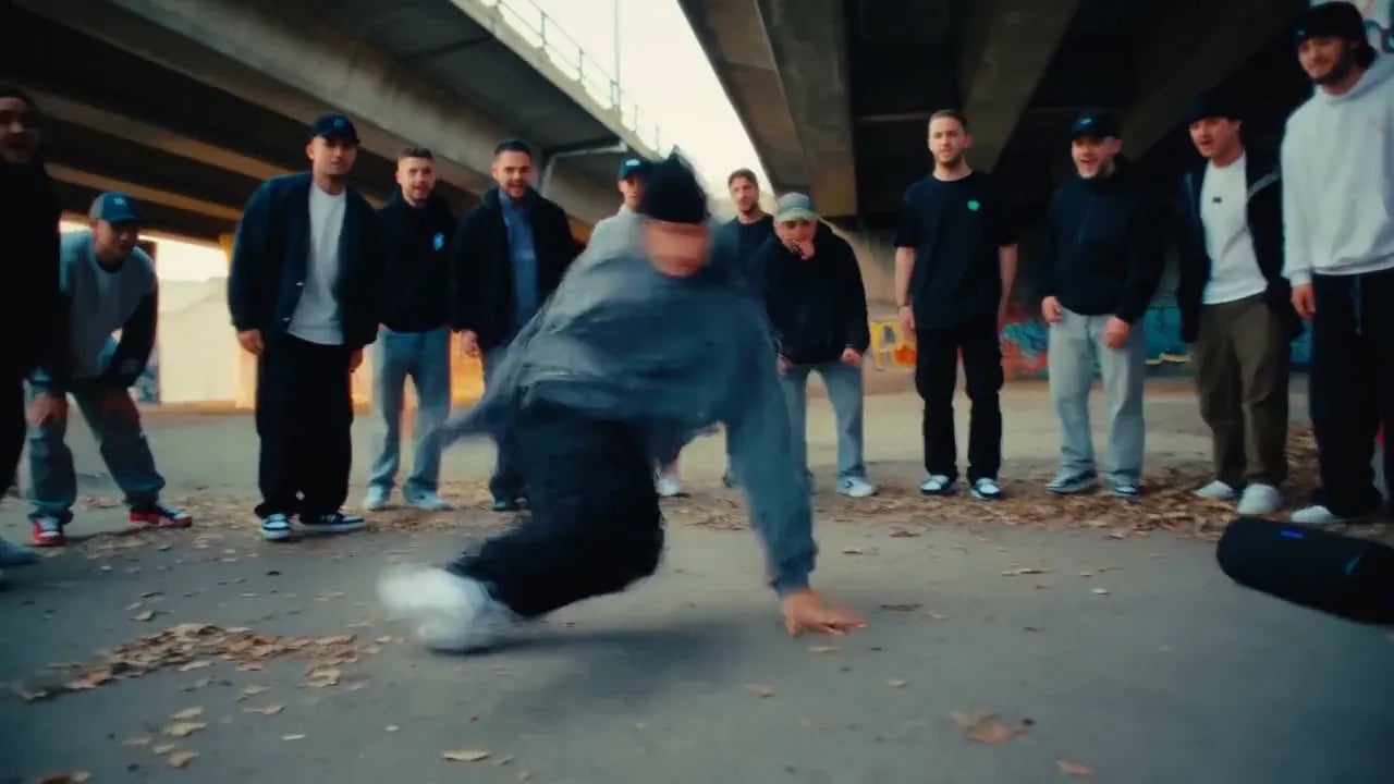 A breakdancer spins on the concrete under a highway overpass while a crowd watches.