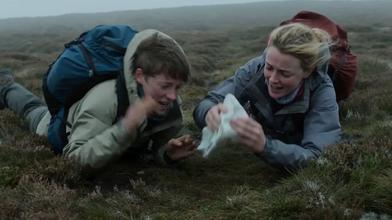 Two hikers lie laughing in the heather as they catch a map on a misty moor.