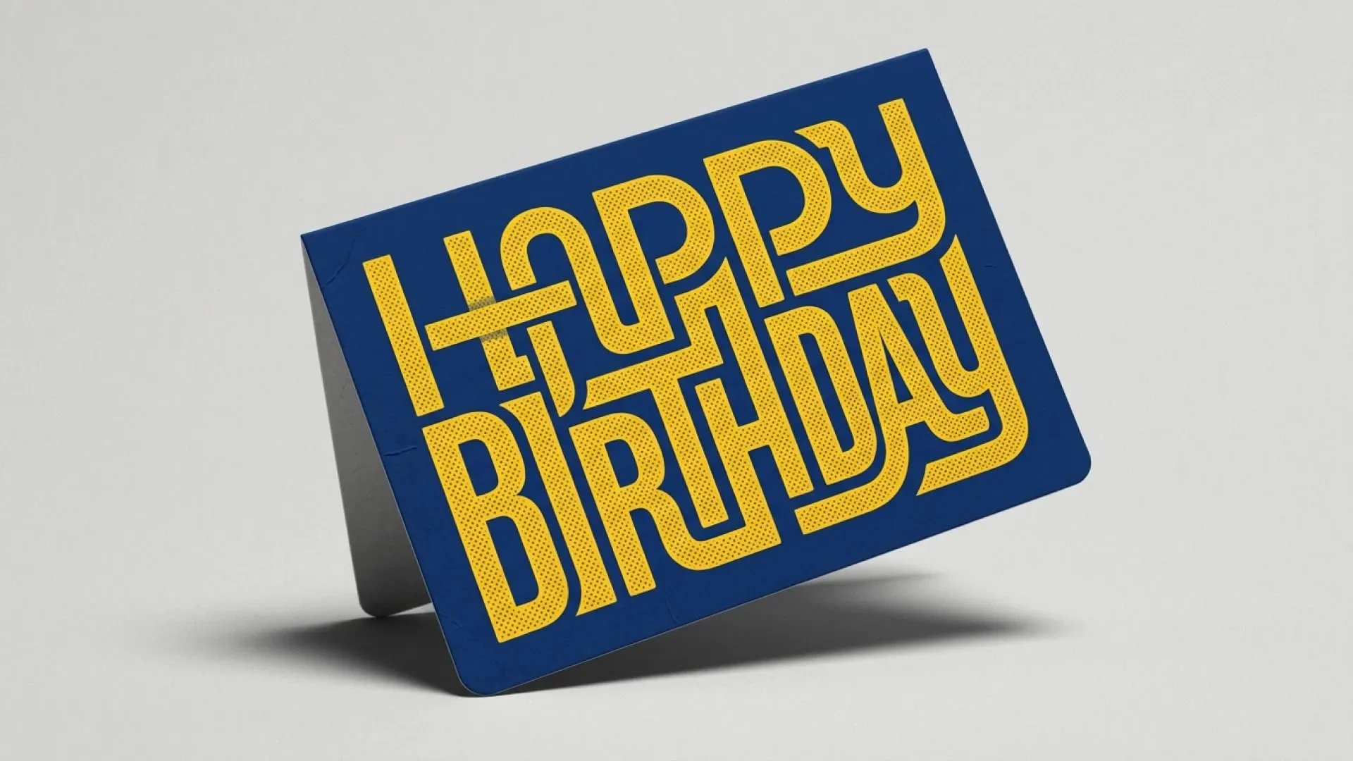 A navy greeting card with interlocking yellow Happy Birthday lettering stands on a light gray surface.