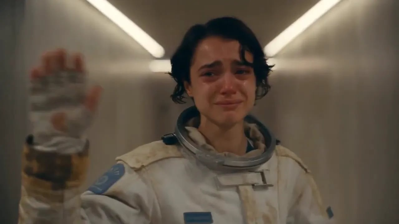 A tearful astronaut in a worn white suit raises a gloved hand in a bright corridor.