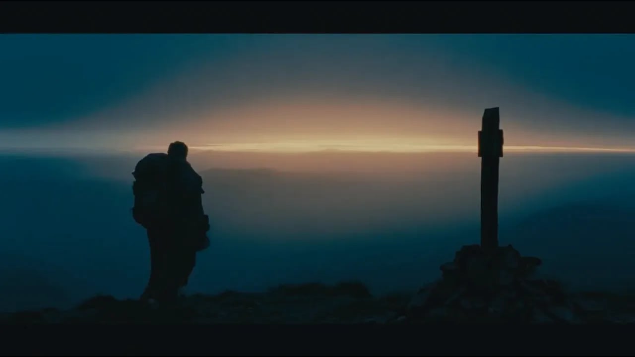 A lone traveler stands in silhouette beside a stone marker at dusk.