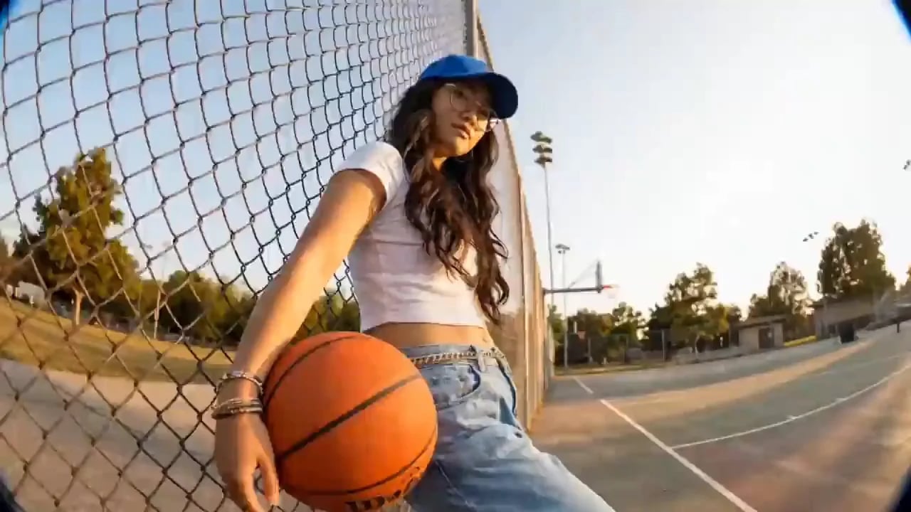 A young woman in a blue cap dribbles a basketball on an outdoor court at sunset.